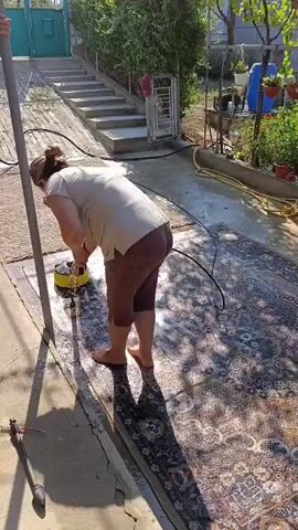 A person is cleaning a patterned rug outdoors. They are using a yellow cleaning machine with a rotating brush. The rug is laid out on a concrete surface. Water is spraying from a hose onto the rug, and the cleaning machine is creating foam. The person is wearing a light-colored t-shirt and brown leggings, and is barefoot. There are stairs and greenery in the background.