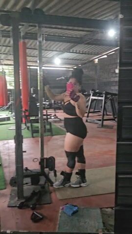 Here's a description of the video:

The video shows the interior of a gym with several people exercising. The floor is a combination of green turf and red concrete. The gym has a partially open roof structure.

A woman wearing a black sports bra, black shorts, knee pads, and gloves is standing near a cable machine. She is holding a pink phone, seemingly taking a photo or video of herself.

In the background, a man wearing a red shirt and black shorts is walking near some dumbbells and exercise equipment. Other people are seen in the background using different equipment, including a woman near a weightlifting machine. There's also a red punching bag visible.