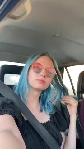 The media shows a close-up shot of a young woman with blue hair, wearing round, ombre sunglasses and a black top. She is sitting in the passenger seat of a car, with a seatbelt fastened across her chest. Her left hand is resting on her lap, and her right hand is near her face, with a ring on her finger. The interior of the car is visible, including the headrest and ceiling. In the background, a man is visible in the driver's seat, wearing a maroon t-shirt and sunglasses. The lighting suggests it is daytime, possibly late afternoon given the warm tones and shadows. The scene appears to be on a road, with glimpses of trees and buildings through the car windows. The overall mood seems relaxed and casual, as if they are on a road trip or driving somewhere pleasant. There is no visible text in the media. The location context provided is "Түндүк айланып өтүүчү жол 84 га, Kyrgyzstan", which translates to "Northern Bypass Road 84, Kyrgyzstan".