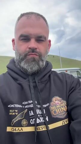 The video begins with a close-up of a man with a beard and a black hoodie, looking directly at the camera. He is outdoors, with a cloudy sky and green hills in the background. As the video progresses, a woman appears next to him, smiling and waving. They appear to be on a boat, with a body of water and distant hills visible. Another person is partially visible in the background. Seagulls are seen flying around. The man and woman are looking at the camera and smiling, suggesting they are taking a selfie video. The woman is wearing a white jacket. The man's hoodie has graphics on it, including text and a circular emblem. The overall scene is a casual, outdoor setting.