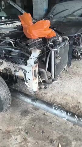 The video shows a white car that has been heavily damaged, possibly in an accident. The front end of the car is disassembled, revealing the engine and other internal components. The hood and bumper are missing, and the front fender is detached. The windshield is cracked and covered with a yellow plastic sheet. The car is in what appears to be a workshop or garage, with a concrete floor and other vehicles or parts visible in the background. There are also some orange and yellow plastic bags draped over parts of the car. The car is supported by a metal rod or pipe that extends from underneath it. The overall impression is of a vehicle undergoing repair or salvage.