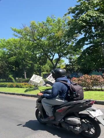A person wearing a helmet and backpack rides a dark-colored scooter on a paved road. The scooter has a white license plate. The person is facing away from the camera and appears to be moving to the right. The background features lush green trees, a grassy area, and a geometric sculpture. The sky is clear and blue. The lighting suggests it is daytime, likely sunny. The scene appears to be an outdoor urban or suburban environment.