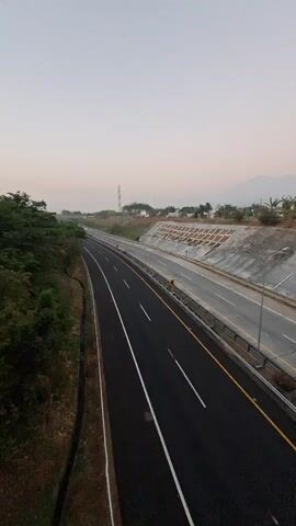 The video shows an aerial view of an empty highway in the early morning. The sky is a pale pink, indicating sunrise. The highway has multiple lanes and is bordered by trees and a retaining wall. There is a single red car driving on the highway. The scene is peaceful and quiet.