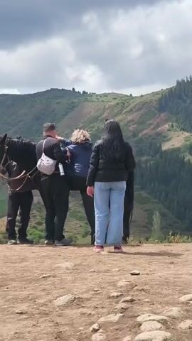 The video captures a group of people in a mountainous outdoor setting in Jeti-Ögüz, Kyrgyzstan. The central activity involves a person with blonde, curly hair, dressed in a blue jacket, being assisted onto a black horse. Two individuals are actively helping: a man in a black outfit with a white crossbody bag supports the rider, while a woman with long dark hair, wearing a black jacket and light blue jeans, stands by the horse's side, also offering support. In the foreground, another person is seen from behind, wearing a pink t-shirt with a graphic of two cartoon characters and the partially visible text "LIFE HAS NO [missing word] GO WILD OUTDOORS!". This person is holding a smartphone, apparently filming the scene. The ground beneath them is a dirt path, suggesting a trail or viewing point. The background reveals a beautiful landscape of green, forested mountains under a cloudy sky. The scene depicts a casual, recreational outdoor activity, likely preparing for a horseback ride in the scenic mountain environment. The time of day appears to be daytime, with natural light under the overcast conditions.