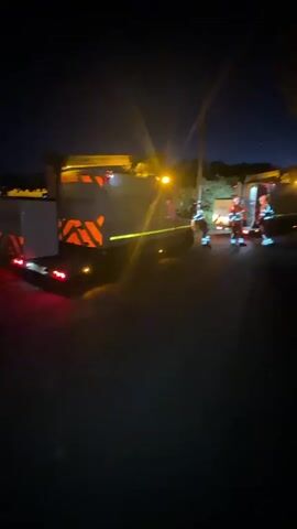 The video depicts a nighttime scene involving multiple utility vehicles and personnel in Babergh, United Kingdom.

In the foreground and to the left, a white work truck or utility vehicle is parked, facing away from the camera. Its rear is prominently displayed, featuring bright red brake lights and distinctive orange and white chevron reflective markings. A yellow crane or boom arm is extended upwards and to the right from the vehicle, equipped with very bright yellow work lights that illuminate the immediate area and create significant lens flares.

Another white vehicle, possibly a van or truck, is visible to the right, also with its rear facing the camera and adorned with similar orange and white chevron markings and bright lights.

Between these two vehicles, several individuals, estimated to be at least four or five, are present. They are dressed in high-visibility work attire, likely orange or red uniforms with reflective stripes, indicating they are working or responding to an incident. A bright yellow or green reflective tape or line is stretched across the ground, possibly delineating a work zone or hazard area.

The entire scene is enveloped in darkness, characteristic of nighttime, with the sky appearing dark blue or black. The primary illumination comes from the powerful work lights mounted on the vehicles, casting stark light and shadows. The background shows dark silhouettes of trees or bushes and a tall, slender pole, possibly a utility pole. The ground appears to be a dark paved surface.

The activity suggests an active work site or an emergency response operation, likely involving maintenance, repair, or an incident requiring specialized vehicles and personnel during the night. No specific text is clearly legible on the vehicles due to the lighting and distance, apart from the chevron patterns.