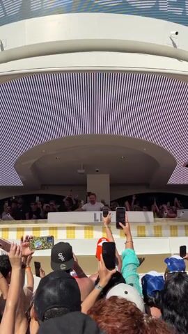 The scene captures a lively atmosphere at a daytime event, possibly a beach club or poolside party. A DJ stands with arms raised in a booth above the crowd, acknowledging the audience. People in the crowd are facing the DJ, many recording the event on their smartphones. A large screen behind the DJ displays vertical light patterns. The crowd is diverse, with various hats, hairstyles, and swimwear. The setting appears to be a well-designed entertainment venue with a mix of architectural and modern design elements.