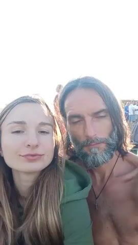 A selfie video shows a couple close together in an outdoor setting with bright sunlight. The woman is on the left, with long, blonde hair and a neutral expression. The man is on the right, with long, dark hair, a beard, and is shirtless, wearing a cross necklace. He is looking at the camera with a slight smile. In the background, other people and structures are visible, suggesting a gathering or event. The sun is very bright, causing some overexposure in the video.