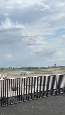 The video shows a Transavia airplane taxiing on a runway at an airport. The airplane is white with green accents and the Transavia logo. It is moving from left to right across the frame. In the foreground, a black metal fence runs horizontally, obscuring the bottom portion of the view. Beyond the fence, the tarmac of the airport is visible, along with a jet bridge on the far left. In the distance, a city skyline can be seen under a cloudy sky. The clouds are a mix of white and grey, suggesting either a partly cloudy day or the possibility of approaching rain. The lighting indicates it is daytime, likely late morning or afternoon. The overall scene is typical of an airport environment, with the focus on the commercial aircraft preparing for or having just completed a flight. There are no people clearly visible in the frame. The text "transavia" is clearly visible on the side of the airplane. The location context provided is Paray-Vieille-Poste, France, which would place this airport as likely Paris Orly Airport (ORY).