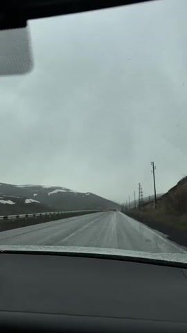 The video, filmed from inside a vehicle, shows a wet, two-lane road stretching into the distance under an overcast sky. The view is through the windshield, with the dashboard visible at the bottom of the frame and a rearview mirror at the top. Windshield wipers are active, clearing raindrops from the glass, indicating rainy or wet weather conditions.

On both sides of the road, there are rolling hills and mountains, with patches of snow visible on some of the higher elevations in the background. A guardrail lines the left side of the road for a portion of the view. To the right, a series of power poles with overhead lines runs parallel to the road. Several vehicles can be seen traveling in the distance, both ahead and approaching. The overall atmosphere is one of a cold, wet day in a mountainous region, consistent with the provided location context of Tsovagyugh, Armenia.