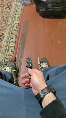 A person wearing blue sweatpants, a black long-sleeved shirt, and a black watch on their left wrist is holding and flipping a folding knife. They are sitting in what appears to be a room with a red linoleum floor. There is a rug with a colorful pattern along the left side of the frame.  Bottles of water, a black garbage bag, and a green trash bin are visible in the background.  The person's feet, shod in sneakers, are also in view.  The camera appears to be positioned from a first-person perspective, looking down at the person's lap.