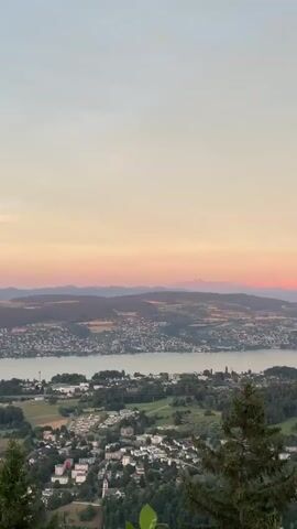 A wide aerial view captures a sunset over a lake and a city. The sky is a gradient of pale yellow to pink, with a few clouds reflecting the sunset's colors. The lake stretches across the middle ground, with a city built along its shores and hills in the background. The foreground shows a portion of a hillside with trees and some buildings. The overall impression is one of natural beauty and urban development.
