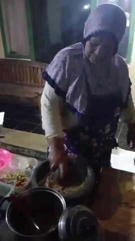 A woman wearing a headscarf and apron is shown from the waist up, standing behind a table. She is actively engaged in preparing food. In front of her, a mortar and pestle are visible, containing ground ingredients. She uses a spoon to stir something in a pot on the table. Various containers of ingredients, including what appear to be chilies and other vegetables, are arranged around the work area. The background suggests an outdoor or semi-outdoor setting, possibly a food stall, with a wooden bench and window visible. The lighting is dim, indicating it might be evening or nighttime.