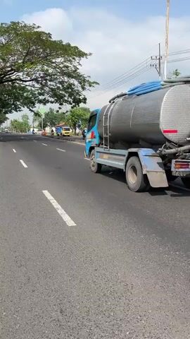A blue tanker truck is driving on a paved road under a partly cloudy sky. The truck has a large silver tank. The road has white lane markings. On the left side of the road, there is a large tree and some greenery. In the background, there are other vehicles and buildings. The camera is at eye level with the truck and is panning slightly to the right.