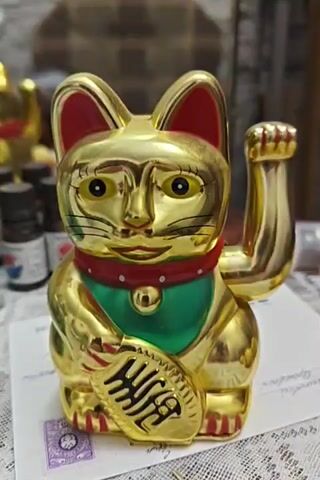 A gold-colored Maneki-neko figurine with a waving paw is centered in the frame. The figurine has a red collar with a green bib and holds a gold ingot in its left paw. The background is blurred, showing indistinct objects and a patterned surface. The lighting is soft, and the focus is on the figurine.