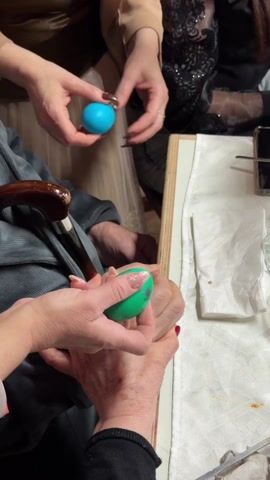 Several individuals are engaged in what appears to be an Easter egg cracking game. One person, wearing a tan blazer, holds a blue egg while another person holds an orange egg. They are gathered around a table covered with a white tablecloth, along with other colored eggs, including green. An older individual with a walking stick is also involved in the game, holding a green egg. Laughter can be heard in the background.