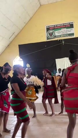 This video depicts a vibrant cultural performance taking place indoors. A group of young women, dressed in traditional attire consisting of red and green patterned wraps around their waists and heads, are engaged in dancing. Their movements are lively and energetic, suggesting a celebratory or festive occasion. One of the dancers in the foreground is wearing a black top with her wrap. Another dancer, further back, has a red cloth draped over her shoulder. In the background, a few individuals are seated, appearing to be an audience. One person in the very back is wearing a white and yellow patterned outfit.

A banner hangs prominently on the wall behind the performers. The text on the banner reads: "FELLOWSHIP OF CHRISTIAN NURSES (FCN) NEXT GENERATION (NG)". Below this, it states: "MOTTO: CARING FOR THE TOTAL MAN; SPIRIT, SOUL AND BODY." Further text indicates "1 Thess. 5:23-24" and "VOM BRANCH 2018/2019". The presence of this banner suggests the event is organized by the Fellowship of Christian Nurses, Vom Branch, for their Next Generation group, likely in 2018 or 2019.

The setting appears to be a hall with a sloped ceiling and wooden beams. The walls are painted yellow and white. The floor is tiled with a light-colored material. Natural light is entering from the left side of the frame, suggesting it might be daytime. The atmosphere seems joyful and engaging, with the dancers clearly immersed in their performance.
