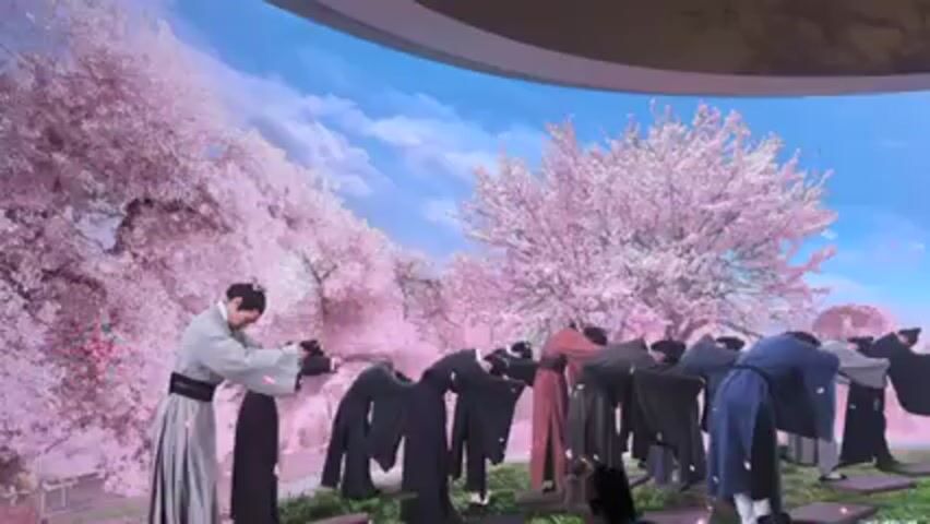 In front of a curved screen displaying a landscape of pink cherry blossom trees under a blue sky, a group of about a dozen people dressed in what appear to be historical East Asian clothing perform a synchronized bow. They stand in a line and move in unison, their clothes varying in color from gray to black to red and blue.  As they straighten up, they look towards the viewer. The location appears to be indoors.