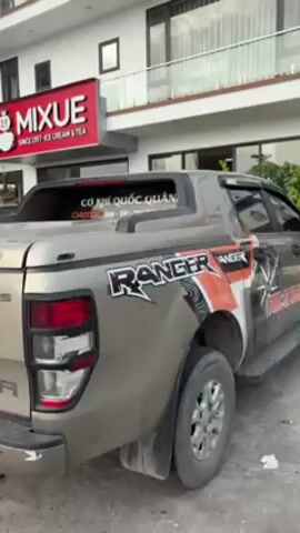 The camera pans along the passenger side of a khaki-colored Ford Ranger pickup truck, heavily decorated with graphics, parked in front of a MIXUE ice cream and tea shop. The truck has a partial bed cover and running boards. The MIXUE sign is bright red and features a cartoon snowman. The building behind the shop is white with multiple windows and balconies.