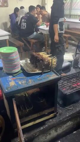 The video shows an outdoor food stall or restaurant setting at night. In the foreground, a barbecue grill with glowing coals is visible, positioned on a stone counter. To the left of the grill is a blue metal cabinet with an open compartment containing food skewers and a pot. A stack of green and white plates sits on a blue surface to the left of the cabinet. A person wearing a black shirt and pants is standing near the grill, appearing to be tending to it. In the background, several people are seated at tables, eating and socializing. One person is wearing a hijab. The environment appears to be somewhat dimly lit, suggesting it is nighttime. There is a blue electric fan in the middle ground. A sign with Indonesian text is visible behind the seated patrons.