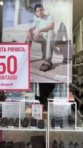 This media file captures a view of a shoe store window display, likely in Livorno, Italy, given the textual cues. The primary focus is on a large advertisement featuring a man wearing Bata sandals, promoting a "Private Sale" with discounts of "50%" and other percentages visible. The background of the ad shows a modern, architectural setting, possibly a staircase.

Below the advertisement, the store's merchandise is displayed in glass cases. One shelf showcases various men's and women's footwear, including loafers, sneakers, and sandals, with price tags of €39.99 and €32.90. Another shelf displays more casual shoes, such as flip-flops and slip-ons, in muted colors. Further down, a selection of athletic shoes and sneakers are visible, some with price tags in the €39.99 range.

The overall scene suggests an indoor retail environment with bright, artificial lighting, characteristic of a store interior. The display is well-organized, aimed at attracting customers with sale promotions and a variety of footwear options. The reflection on the glass suggests it is daytime, and the overall atmosphere appears to be that of a typical commercial street. The visible text, "VENTA PRIVATA," "20 30 40 50," and "Bata MAKE YOUR WAY," clearly indicates a sale event and the brand name.