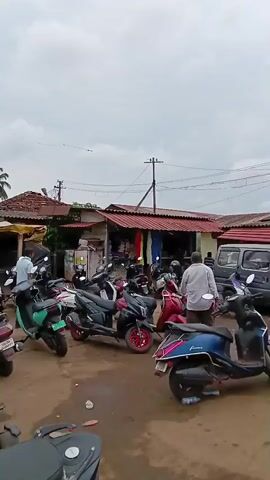 The video depicts a street scene in what appears to be a small town, possibly in India. The weather seems overcast. A large number of scooters and motorbikes are parked haphazardly in a dirt lot. Several small shops and buildings line the street, some with awnings or covered areas. A few people are visible, including adults and children, walking through the area and browsing the shops. Palm trees are seen in the background. There are power lines visible, strung between poles. The overall impression is a bustling, local market or commercial area in a tropical or subtropical climate.