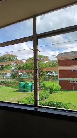 The video shows an interior view looking out a window. The window has horizontal glass slats that are partially open. Outside, there is a grassy field with trees in the background. A brick building is visible on the right side of the frame. There are a few green water tanks in the middle of the field. The sky is blue with some clouds. The inside of the room where the video is taken appears dark.