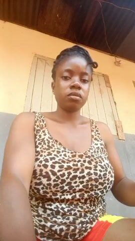 A woman is sitting in front of a beige building with a white and gray window behind her. The ceiling is a brown corrugated metal structure. She is wearing a leopard print top and red shorts. She briefly touches her hair, which is styled in a bun. The woman has dark skin and a serious expression.