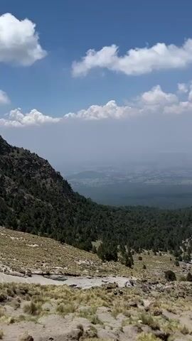 The video depicts a scenic view from a mountain overlooking a valley, partially obscured by haze. The foreground features a rocky, grassy slope with scattered trees. In the middle ground, a dense forest stretches towards the valley. Beyond the forest, a vast, hazy valley floor with signs of urban development is visible under a bright, blue sky dotted with fluffy white clouds.  The camera slowly pans from left to right, revealing more of the expansive landscape.  The environment appears natural with minimal human intervention on the immediate mountainside.