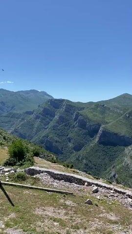 A vertical, outdoor, daytime, landscape video pans slowly across a mountainous, verdant, and rugged terrain under a clear blue sky. In the foreground, a crumbling stone wall borders a grassy area with scattered rocks and a wooden post. Beyond the wall, a steep mountain slope covered in lush green trees and vegetation descends into a deep valley. In the middle ground, a solitary lamppost stands to the right. Further in the distance, layers of progressively lighter green mountains stretch towards the horizon, with a winding road visible in the valley.