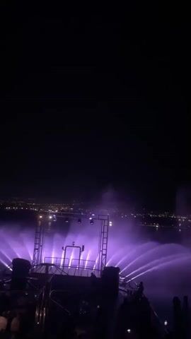 The video shows a nighttime outdoor event with a stage setup and a water fountain show. The stage has lights that cycle through purple, yellow, and green. The water fountains are illuminated by these lights, creating a vibrant display that extends across the background, where a city skyline is visible in the distance. Silhouettes of people are visible in the foreground, suggesting an audience. The overall atmosphere appears to be a festive celebration or performance.