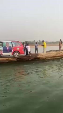 The video shows a river scene, likely in a tropical or semi-tropical region. A car with political posters is on a makeshift wooden ferry. The car is painted in blue, white and red. There are several people on the ferry near the car, some appear to be assisting in the river crossing. The ferry is being propelled across the river by a person in a separate wooden boat, using an oar or pole. The opposite riverbank is sandy with some vegetation and possibly a small village. The sky is hazy and the overall lighting indicates it is daytime.