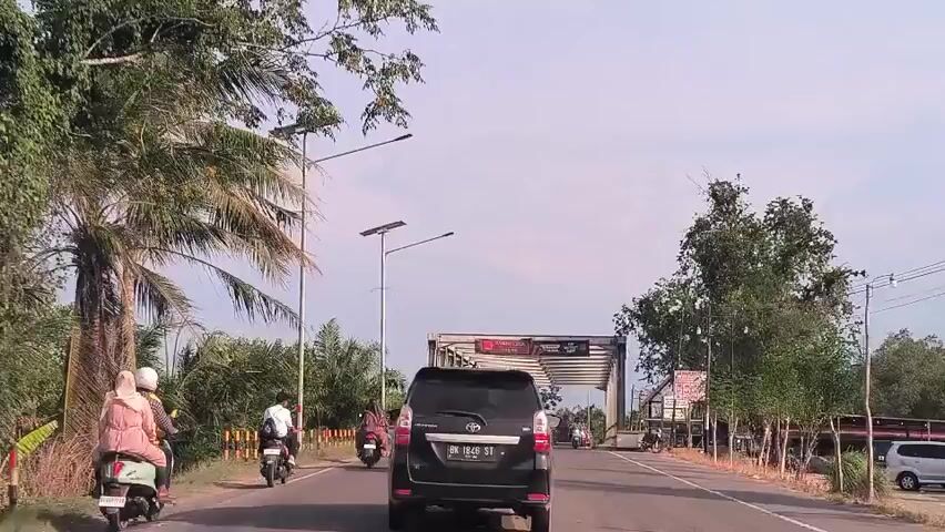 The video shows a car driving down a road toward a bridge. Several motorcycles are also traveling on the road, some ahead of the car and some alongside it. On either side of the road, there are palm trees and other trees. Solar-powered streetlights line the road. There is a building with a red roof to the right of the road. The sky is partially cloudy.