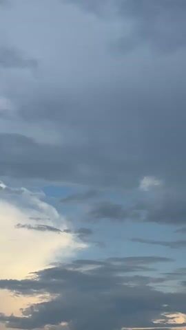 This media file is a vertical video of a cloudy sky, likely captured at sunset or sunrise. The clouds are a mix of dark gray and lighter white and blue, suggesting a dynamic weather system. The lower portion of the frame shows a sliver of pale yellow, indicating the sun is low on the horizon. There are no discernible objects or people in the frame. The scene is natural and evokes a sense of calm or perhaps anticipation of changing weather. The location is identified as Pojarevo, Bulgaria. There is no visible text.