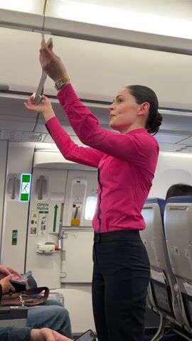 A flight attendant in a pink shirt and black pants stands in an airplane aisle, demonstrating the use of a seatbelt. She holds the seatbelt above her head with both hands. The airplane door with an emergency exit sign is visible in the background. Several passengers are seated, some looking at their phones. The interior of the plane is visible, including seats and overhead compartments.