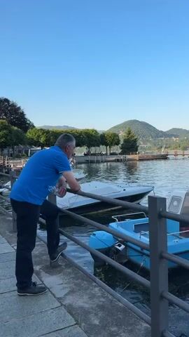 A man in a blue shirt and dark pants leans on a metal fence, looking out at a lake with several boats. A small island is visible in the distance with trees and some buildings. Behind the island are green mountains under a clear, blue sky. The camera pans left to show more boats in the lake. Some of the boats are docked. One is a small sailboat. The water is calm and ripples slightly. The overall tone is peaceful and serene.