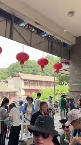 The video shows a crowd of people, mostly dressed in casual summer clothing, queuing in what appears to be a tourist attraction. The environment suggests an outdoor setting with a building made of brick and stone visible, along with lush green trees and foliage. Red lanterns hang from an overhead structure, indicating a possible East Asian influence. The people are standing in a line, some looking at their phones, others talking amongst themselves, suggesting they are waiting for admission or access to a particular area. There are barriers in place to guide the queue. The overall atmosphere seems to be one of orderly waiting during a busy day.