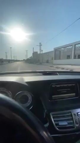 The video, shot from the driver's seat of a car, depicts a drive through a war-torn landscape under a bright sun. The environment is arid, with dilapidated buildings and debris visible on either side of the road. Electrical towers and power lines stand along the roadside. The car proceeds along the road, passing damaged structures and evidence of conflict. The rearview mirror is visible, adding another perspective to the scene.