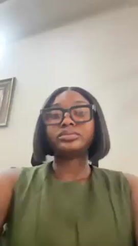 A medium shot shows a woman with dark skin, black rectangular glasses, and a short dark bob haircut. She wears a sleeveless, olive green top. The background is a plain, light-colored wall with a framed picture visible in the upper left corner. The woman is looking directly at the camera.