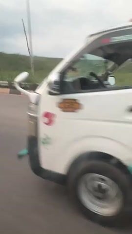A car is driving down a road. The camera is looking out the side window, and the view is of the roadside. There is a hill covered in greenery, a roadside barrier, and an overpass overhead. A flatbed truck parked on the side of the road is seen. There are also a few people standing on the side of the road.