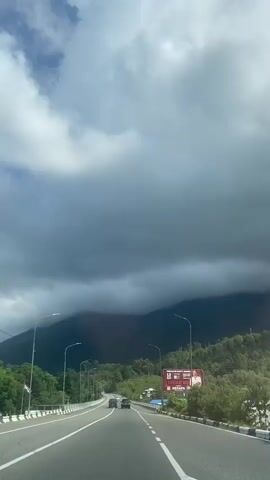 The video shows a view from inside a vehicle driving on a paved road. The road has two lanes in each direction, separated by a white line, and a white and blue barrier on the right side. There are streetlights lining the road. In the distance, there are green mountains covered with trees, and dark clouds are gathering over them. A few cars are visible on the road ahead. There is also a billboard on the right side of the road and a bus stop sign with a speed limit of 90.