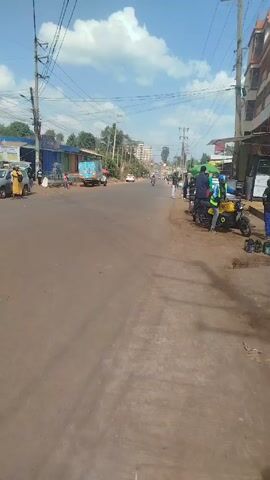 The video shows a street scene in what appears to be a small town or suburb. The road is paved but looks a little worn. There are power lines running overhead, supported by utility poles. Along the side of the road are people standing and walking. There are parked motorcycles. A small blue truck is visible in the distance. Some small shops or stalls are set up along the edge of the road. Other cars and motorcycles pass on the road. The sky is blue with some white clouds.