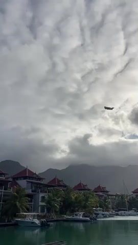 An airplane flies across a cloudy sky over a harbor with boats and buildings. The buildings have red roofs and palm trees in front of them. Mountains are in the background.