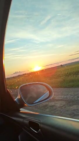 The video shows a view from inside a car, looking out the passenger window. The sun is setting, casting a warm glow over a rural landscape. A field of crops stretches out to the horizon, with a dirt road running alongside it. The side mirror of the car is visible, reflecting a person's arm and hand holding a phone. The overall mood is peaceful and serene, capturing a moment of natural beauty during a car ride.