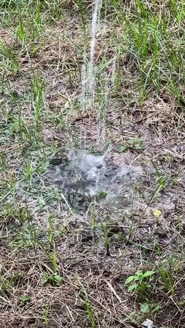 A thin stream of water sprays onto dry grass and dirt from an unseen source above and to the right. The water creates a small, shallow puddle where it hits the ground, splashing outwards and soaking the surrounding dry vegetation. The video focuses on the impact of the water on the ground, showing the dynamic splash and the contrast between the wet and dry elements of the environment.
