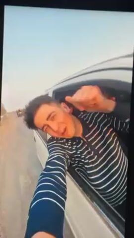 The video shows a young man in the back seat of a white car, leaning out the window. He's wearing a long-sleeved shirt with navy blue and white horizontal stripes. He is looking at the camera and talking while holding the camera up. Other people are visible inside the car. The car is driving on an asphalt road next to a dirt shoulder. Another vehicle, possibly a motorcycle or small car, is visible on the road ahead. The sky is light blue.