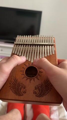 The video shows a close-up of a person's hands playing a kalimba. The kalimba is made of wood and has metal tines. The person is holding the kalimba with their left hand and plucking the tines with their right thumb and index finger. The person is wearing red socks. In the background, a television screen is visible. The video is shot from a first-person perspective.