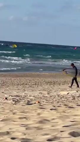 A man in a wetsuit is walking across a sandy beach, carrying a surfboard under his arm. The ocean is to his right, with visible waves and several kitesurfers in the distance. The sky is partly cloudy. There are some discarded objects on the sand in the foreground.