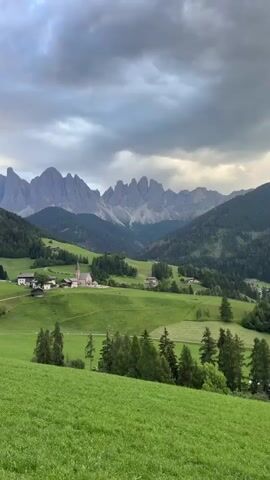 This is a video of a beautiful, scenic landscape. The primary subjects are the rolling green hills and the majestic, jagged mountains in the distance. A small village with a church is nestled in the valley below the mountains. The scene is characterized by lush green fields, dotted with trees and a few scattered houses.

The activity taking place is simply the observation of this natural beauty. There are no people or specific events depicted.

Notable details include the time of day, which appears to be late afternoon or early evening, given the soft lighting and the gathering clouds. The weather seems overcast with a dramatic, cloudy sky, adding to the atmospheric quality of the scene. The location is clearly identifiable as St. Magdalena (Santa Maddalena), Italy, known for its iconic views of the Odle/Geisler mountain range. The emotions evoked are likely those of peace, awe, and tranquility, inspired by the stunning natural panorama.

There is no visible text in the video.