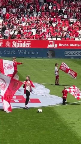 The video shows a soccer game. The stands are full of spectators, the vast majority wearing red. In the foreground, a group of adults and children are waving large red and white flags with the team's logo. A large banner on the field has the team's name written on it. A soccer ball is on the field. A referee runs past.