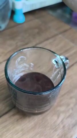 The video shows a close-up of a clear glass mug on a wooden surface. The mug contains a dark liquid, possibly coffee or tea, at the bottom. Hot water is being poured from a transparent pitcher into the mug, creating bubbles and ripples on the surface of the liquid. The water stream is thin and steady, and the pouring action is the primary focus of the short clip. In the background, blurred objects, possibly kitchen items, are visible.